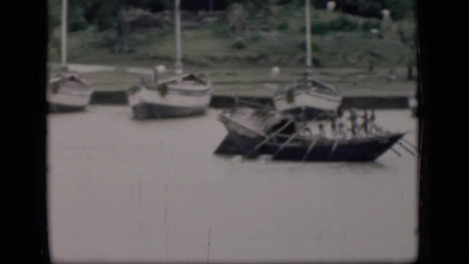 1946 Bombay river view with a traditional wooden boat being rowed by several people, with small sailboats moored behind it.