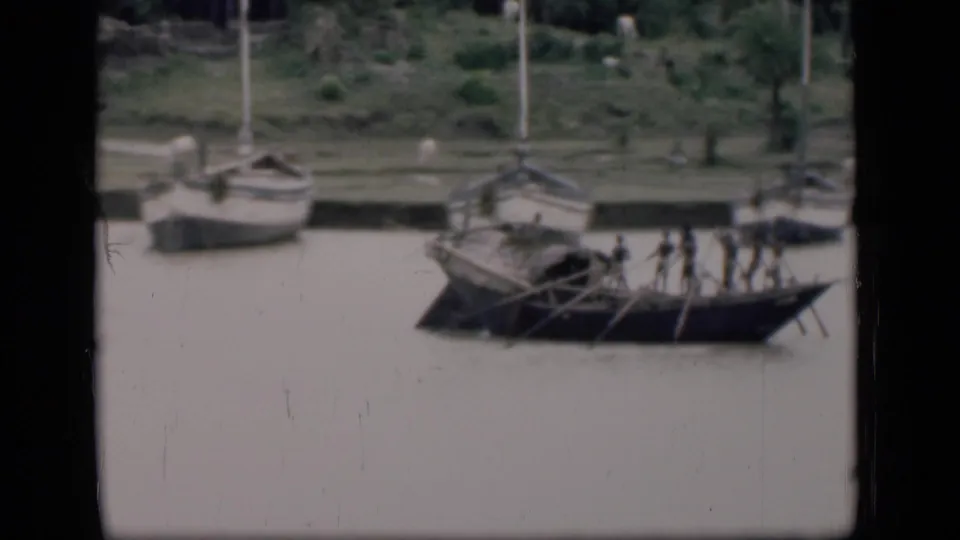 1946 Bombay frame showing a long oared wooden boat on the river, with sailboats pulled close to the grassy bank behind it.