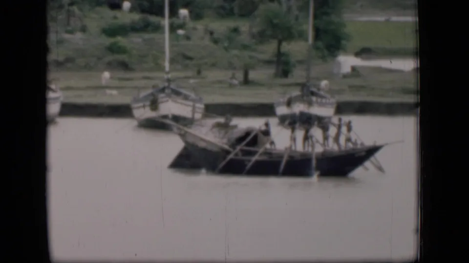 1946 Bombay home-movie frame of several oarsmen in a long wooden river boat, with moored sailboats and shoreline visible behind.