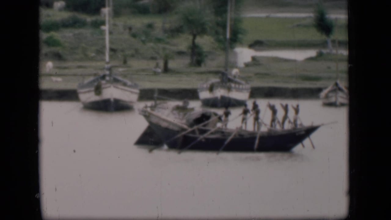 Long wooden boat with standing rowers on a muddy river near a grassy bank.