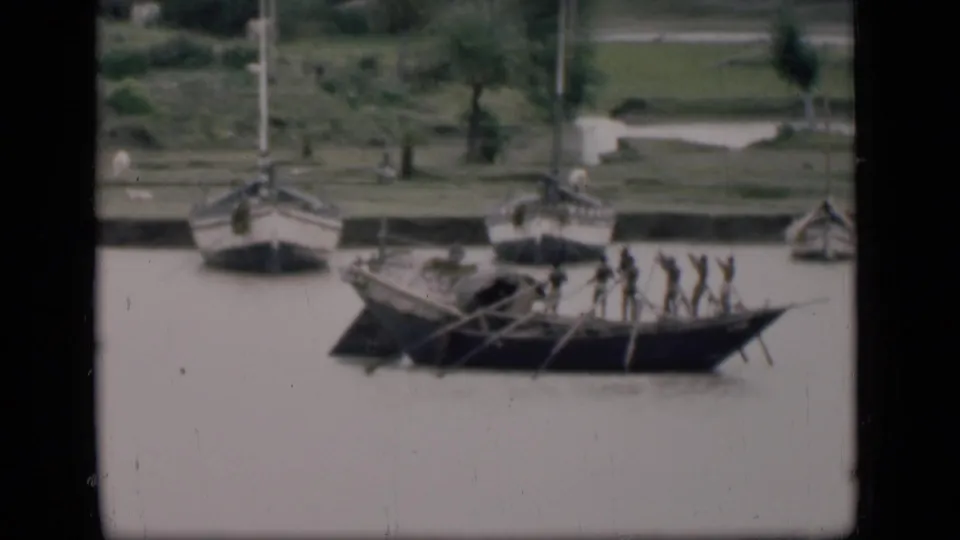 1946 Bombay 8mm frame showing a long wooden boat with several people rowing on a muddy river, with small boats near the bank.