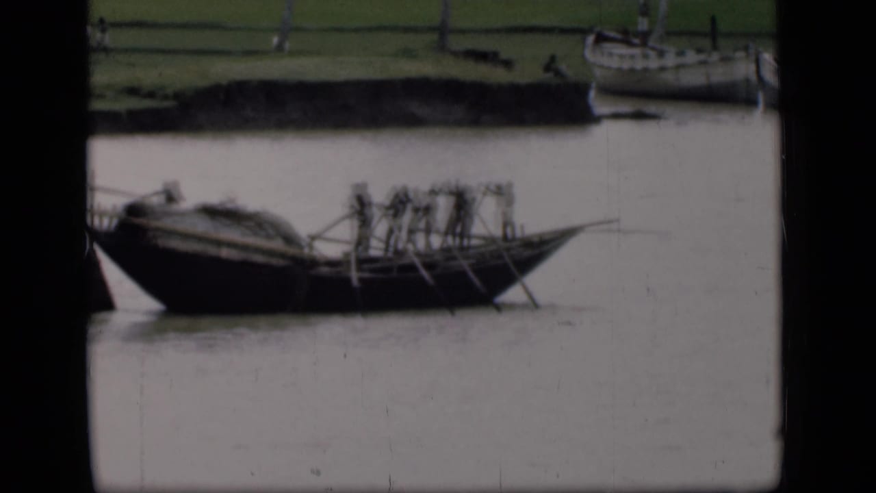 1946 Bombay home-movie frame of a dark wooden boat carrying rowers with long oars on a muddy river beside moored boats.