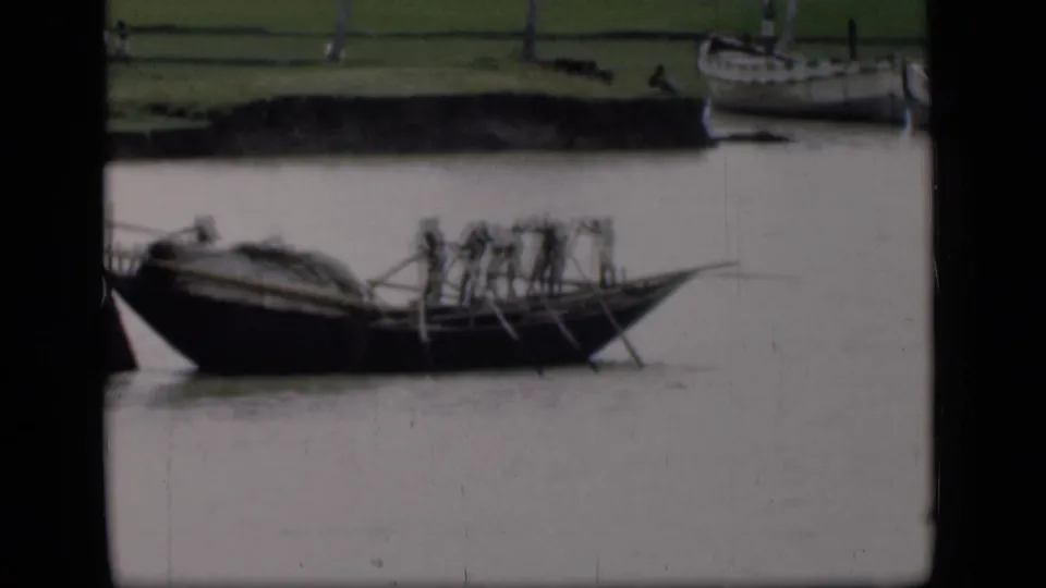1946 Bombay home-movie frame of a dark wooden boat carrying rowers with long oars on a muddy river beside moored boats.