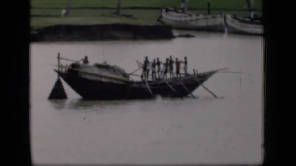 Mid-1940s Bombay 8mm frame showing people rowing a traditional wooden boat on a muddy river with boats along the bank.