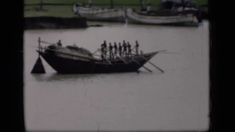 1946 Bombay 8mm frame of a long wooden boat with standing rowers and extended oars, filmed from across a muddy river.