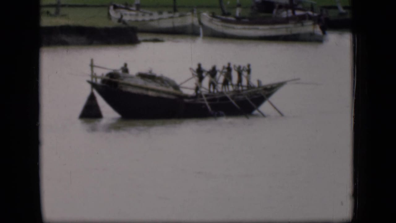 Wooden boat with several people on a calm canal, with other boats partly visible behind it.