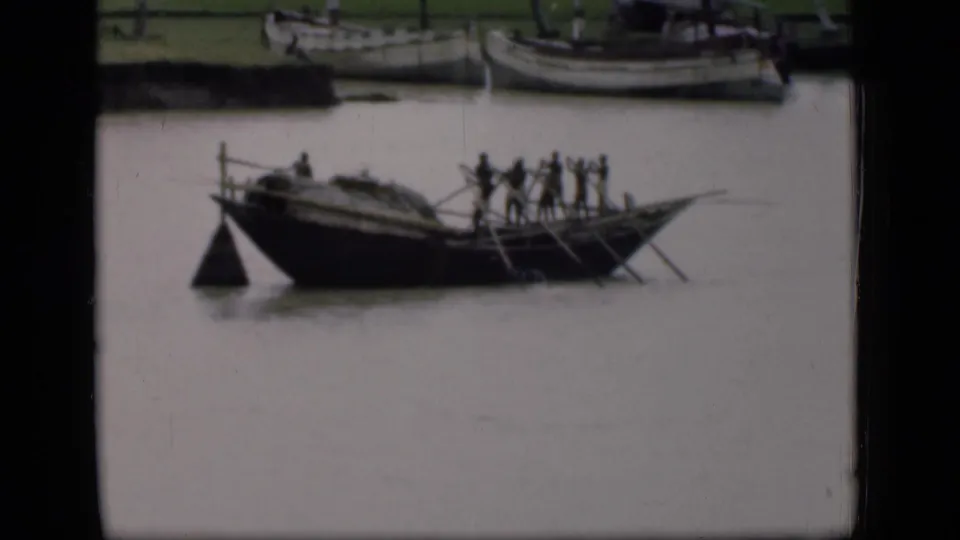 1946 Bombay home-movie frame showing a small wooden boat carrying several people across a canal, with moored boats in the background.