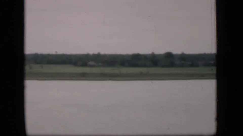 1946 Bombay 8mm frame showing a broad water surface, low shoreline, distant trees, and pale sky from a moving viewpoint.