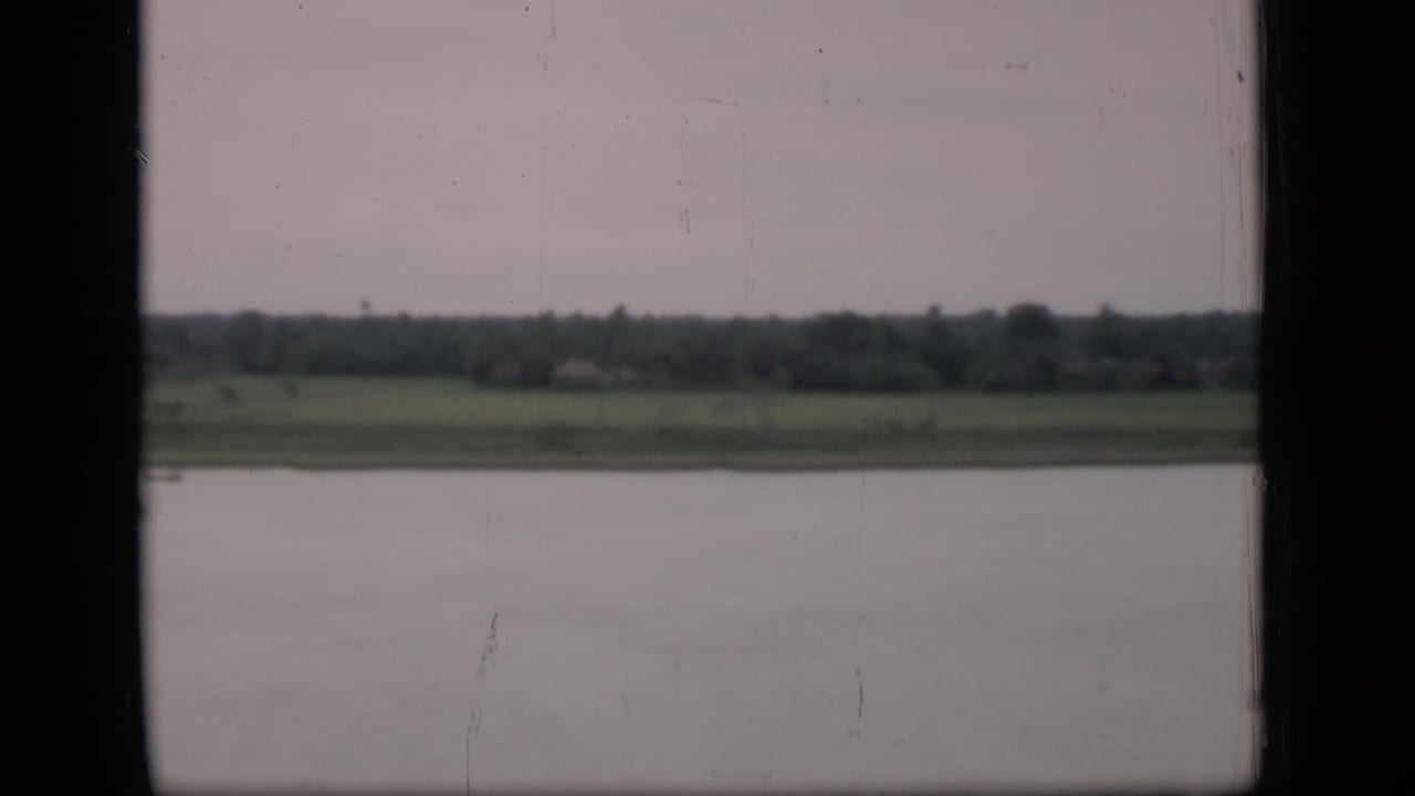 1946 Bombay river view from a boat or window, showing calm water, distant grassy bank, tree line, and overcast sky.