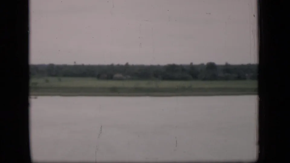 1946 Bombay river view from a boat or window, showing calm water, distant grassy bank, tree line, and overcast sky.