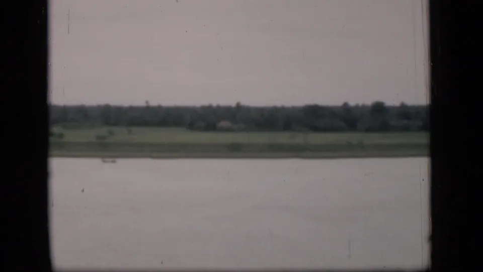 1946 Bombay riverside view with calm water, low green bank, tree-lined horizon, and a small distant boat.