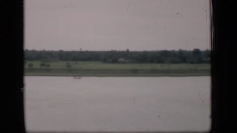1946 Bombay river passage viewed from a moving boat, with water in the foreground, a distant small boat, grassy bank, and trees.