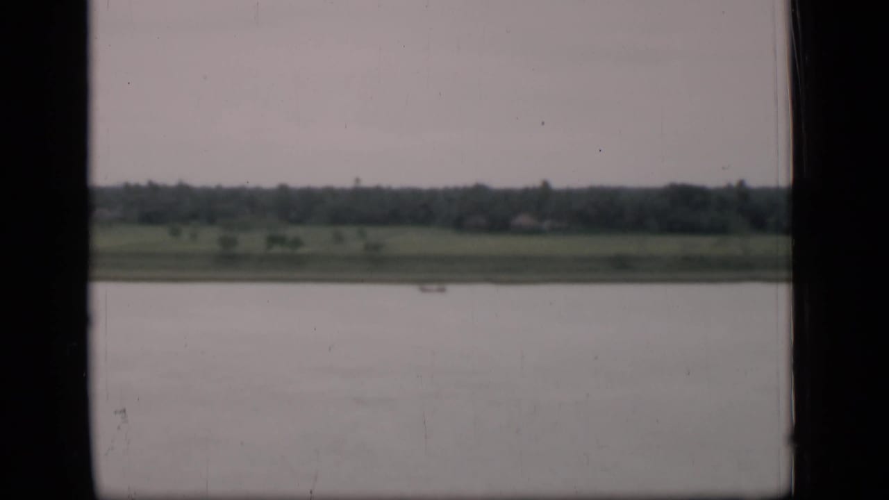 River or lake in foreground with green fields and distant treeline seen through dark window edges.