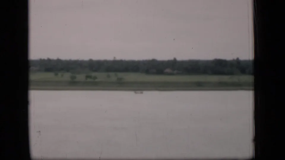 1946 Bombay-area home-movie view across water toward green fields and a distant treeline, framed by dark vehicle or train window edges.