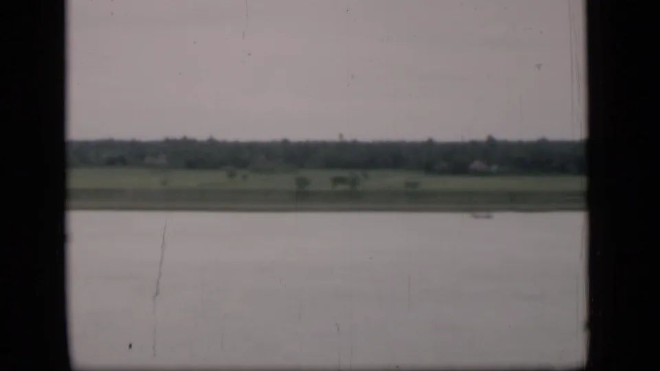 The 8mm frame looks across water toward Bombay-area fields and trees, with a dark central window edge suggesting a moving vehicle viewpoint.