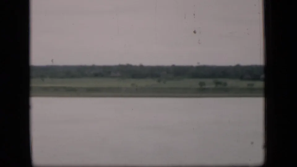1946 Bombay home-movie frame showing a calm waterway and low green shoreline beneath an overcast sky, viewed through dark frame edges.