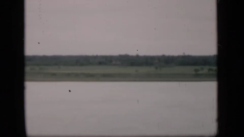 1946 Bombay river view from a moving vessel, showing open water, a low green shore, and dark window-like edges in the frame.
