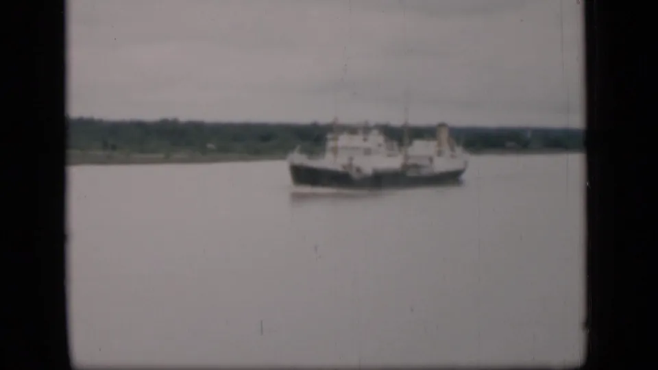1946 Bombay frame with a distant cargo ship crossing calm water, backed by a low green shoreline and cloudy sky.