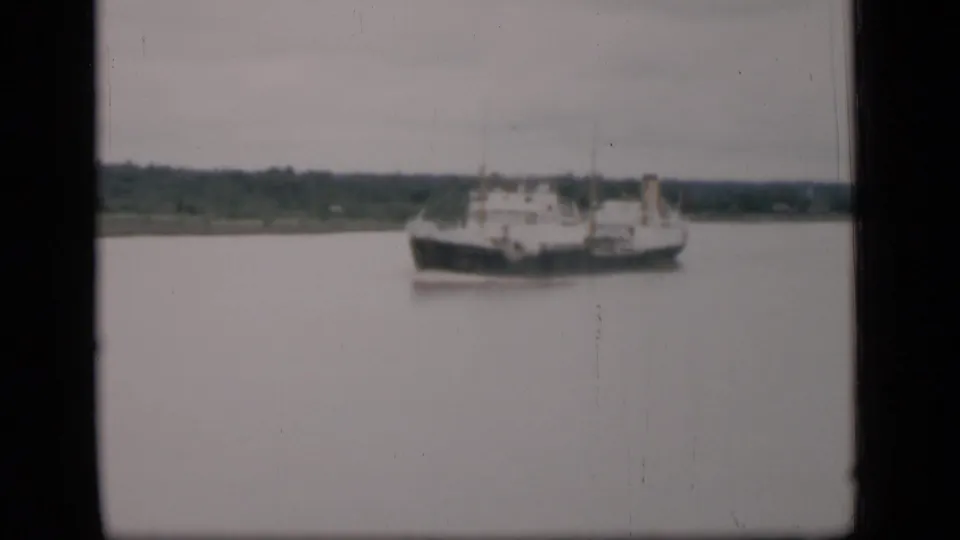 1946 Bombay home-movie still showing a cargo ship on a broad waterway with low green shore in the background.