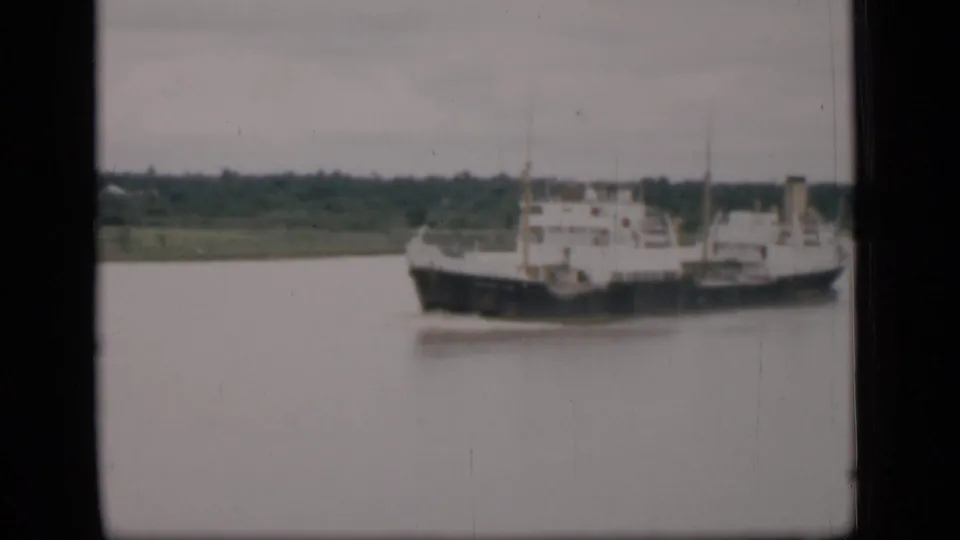 1946 Bombay home-movie frame of a cargo vessel on calm river water, seen from a distant fixed viewpoint.