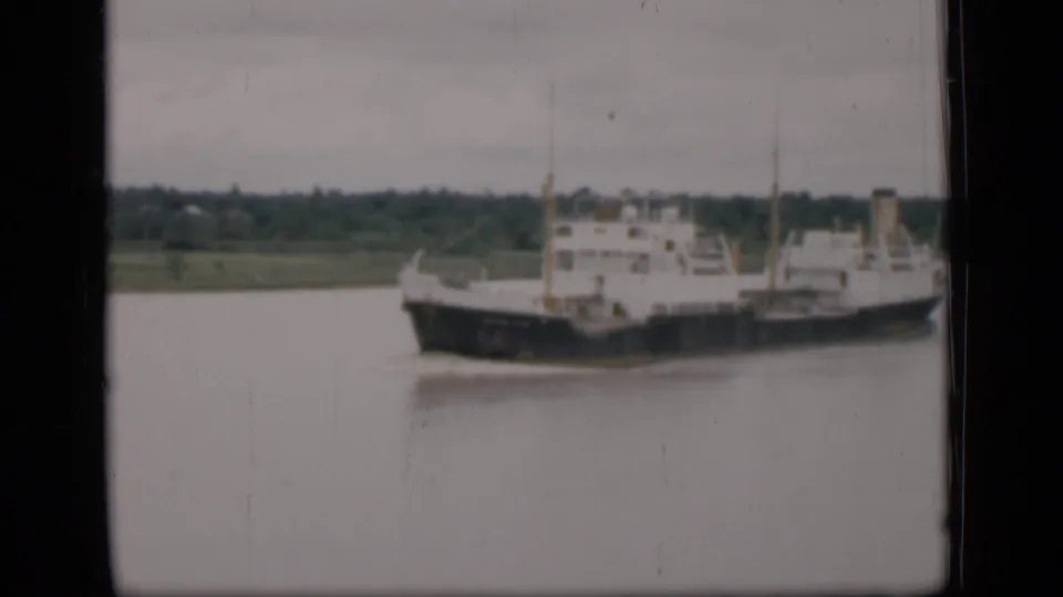 1946 Bombay 8mm frame showing the cargo ship slightly larger against a soft, tree-lined riverbank.