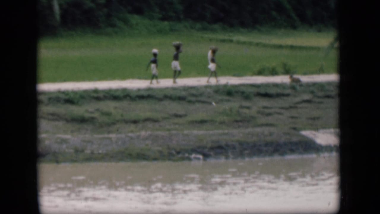 In 1946 Bombay home-movie footage, three distant figures appear to carry loads while walking along a green riverside path.