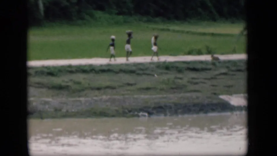 In 1946 Bombay home-movie footage, three distant figures appear to carry loads while walking along a green riverside path.