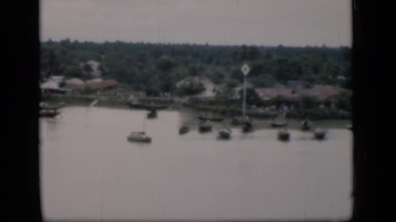 1946 Bombay home-movie frame showing small boats on calm water near low riverside houses and tropical trees.