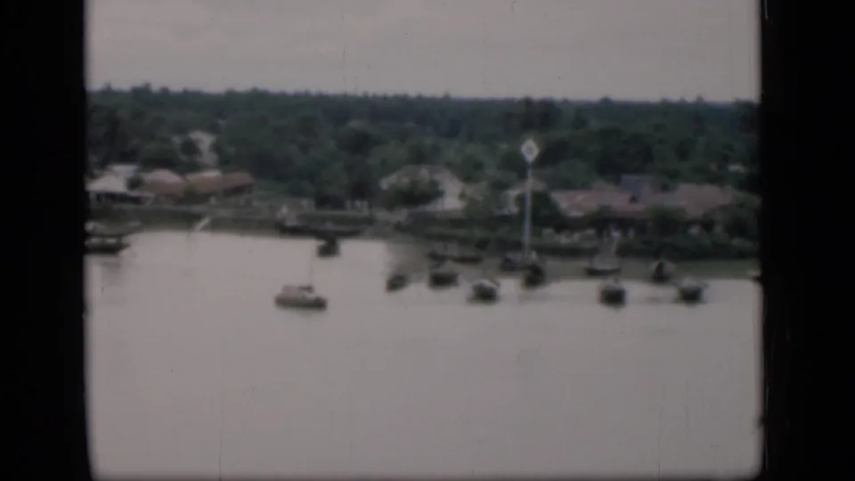 1946 Bombay home-movie frame showing small boats on calm water near low riverside houses and tropical trees.