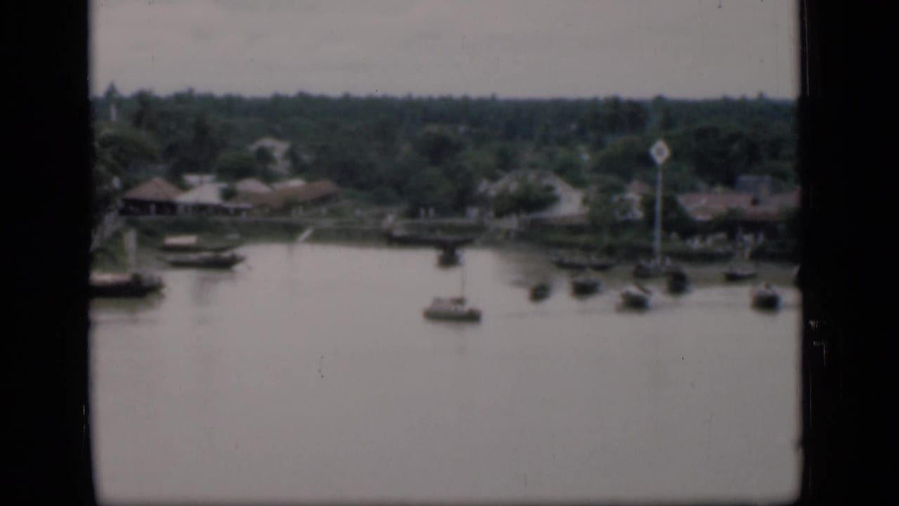 Riverbank with moored boats, low houses, trees, and a tall marker across the water.