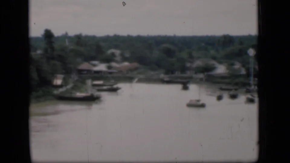 1946 Bombay home-movie frame of a river bend with small boats, tropical trees, and distant waterside buildings.