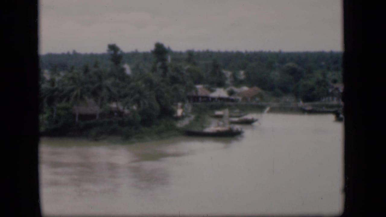 1946 Bombay home-movie frame showing a broad river, palms, moored boats, and low buildings along the far bank.