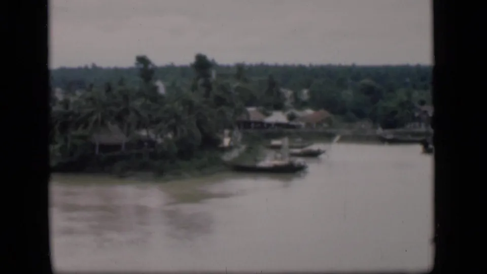 1946 Bombay home-movie frame showing a broad river, palms, moored boats, and low buildings along the far bank.