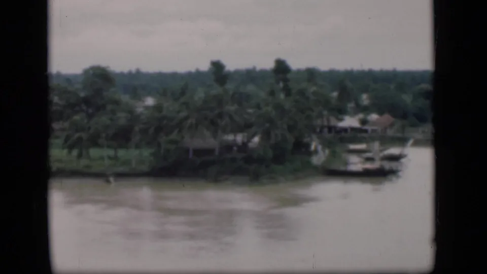 1946 Bombay riverside view with palm trees, small boats near the bank, and low structures under a hazy sky.