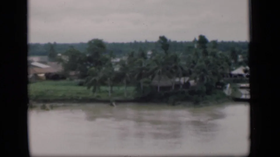 1946 Bombay frame showing a muddy river before a green bank with palms, thatched roofs, and a boat near the shore.