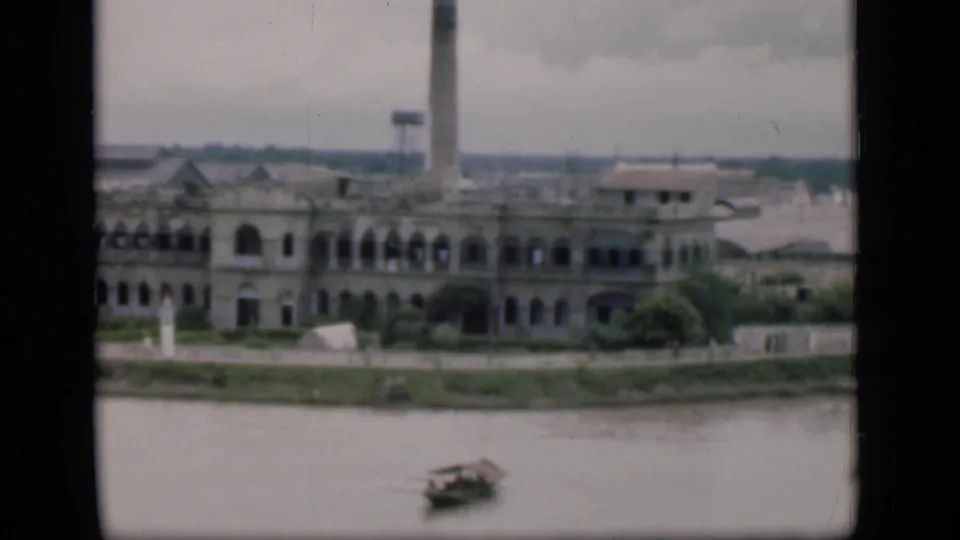 1946 Bombay riverside frame showing a pale multi-story building, arched openings, a tall stack or tower, and a boat crossing the river.