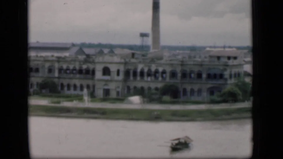 1946 Bombay riverfront view with a large arched building, smokestack, shoreline, and small boat in the foreground.