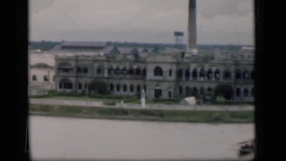 Bombay waterfront pan showing the arched riverfront structure, tall smokestack, open water, and a small boat at the lower right.