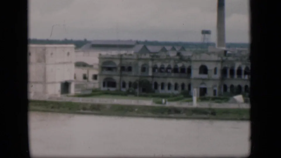 1946 Bombay riverfront view during the pan, with a pale adjoining wall, arched building section, smokestack, and water below.