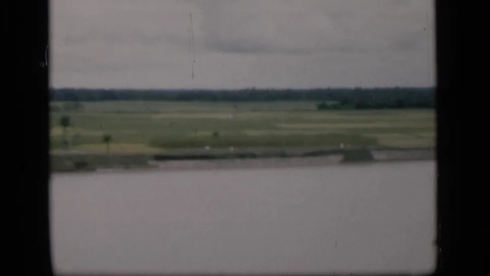 1946 Bombay home-movie frame showing pale water, a low shoreline, green fields, and distant trees under overcast light.