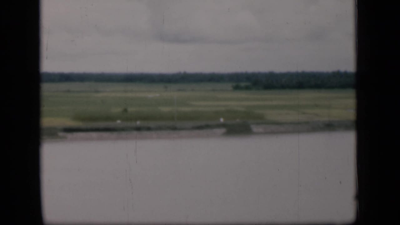 1946 Bombay frame with a broad water foreground, marshy green fields beyond, and a low tree line beneath a cloudy sky.