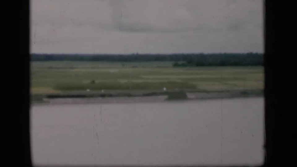 1946 Bombay frame with a broad water foreground, marshy green fields beyond, and a low tree line beneath a cloudy sky.