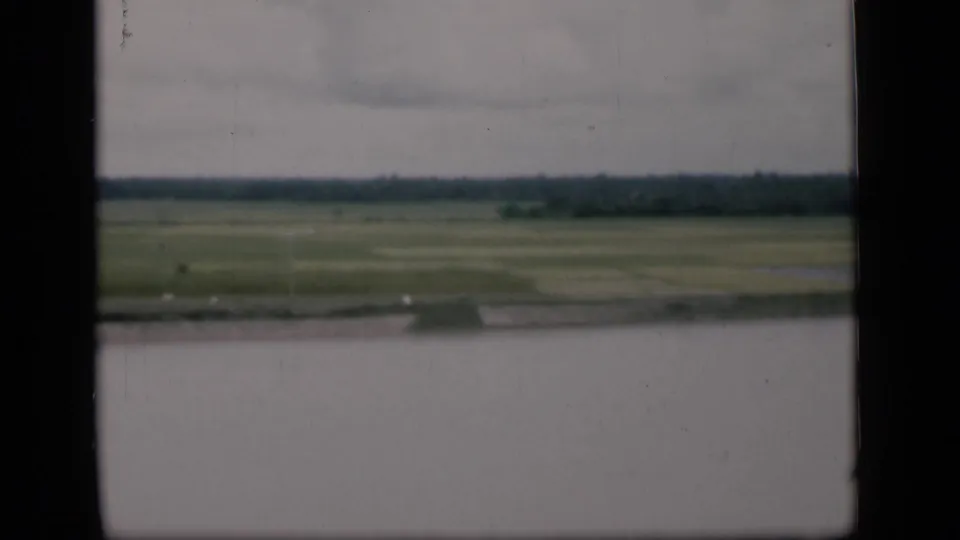 Moving 8mm view from 1946 Bombay showing a waterside bank, open green fields, distant tree line, and visible film wear.