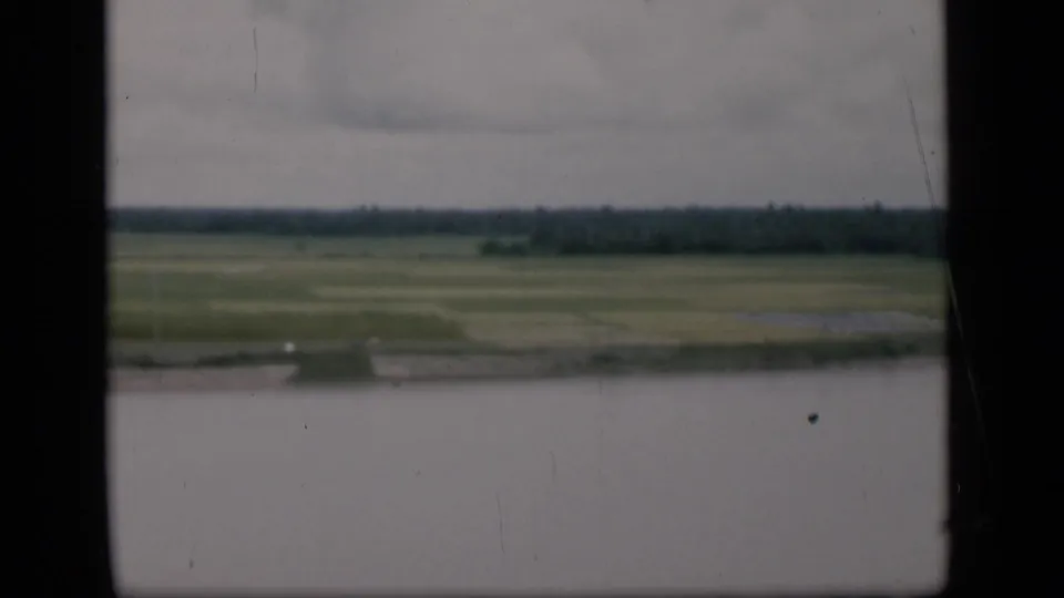 1946 Bombay home-movie still with water in the foreground, green fields beyond, a distant tree line, and muted overcast light.