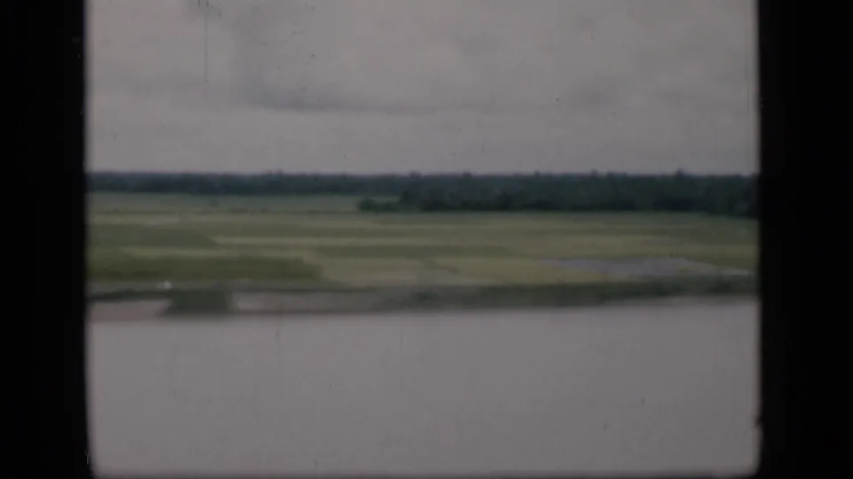 1946 Bombay home-movie view from the water toward flat green riverside fields and a dark tree line beneath cloudy sky.