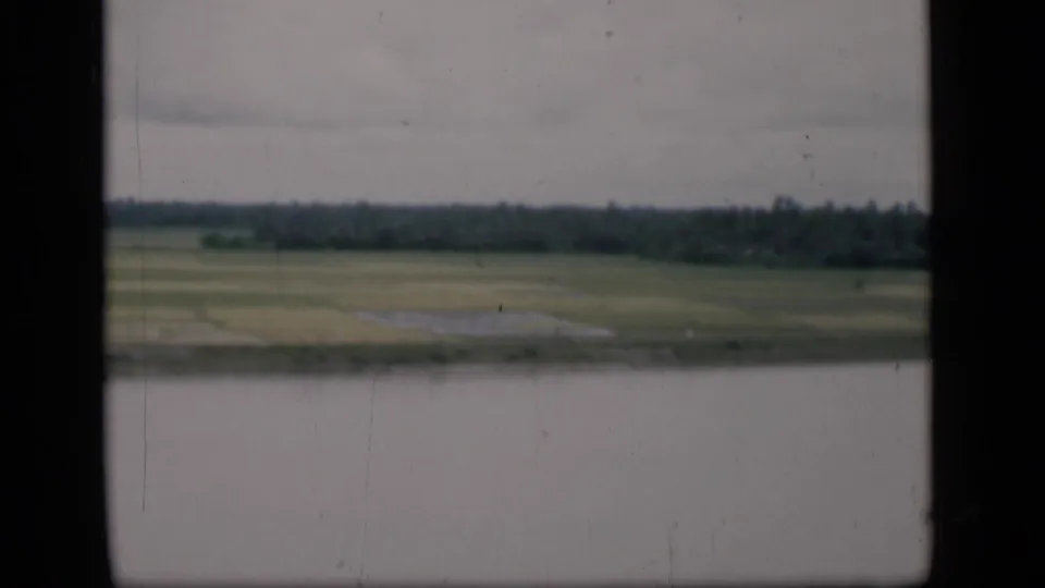 A soft 8mm frame shows Bombay riverside fields across still water, with a dark tree line and cloudy sky in the distance.