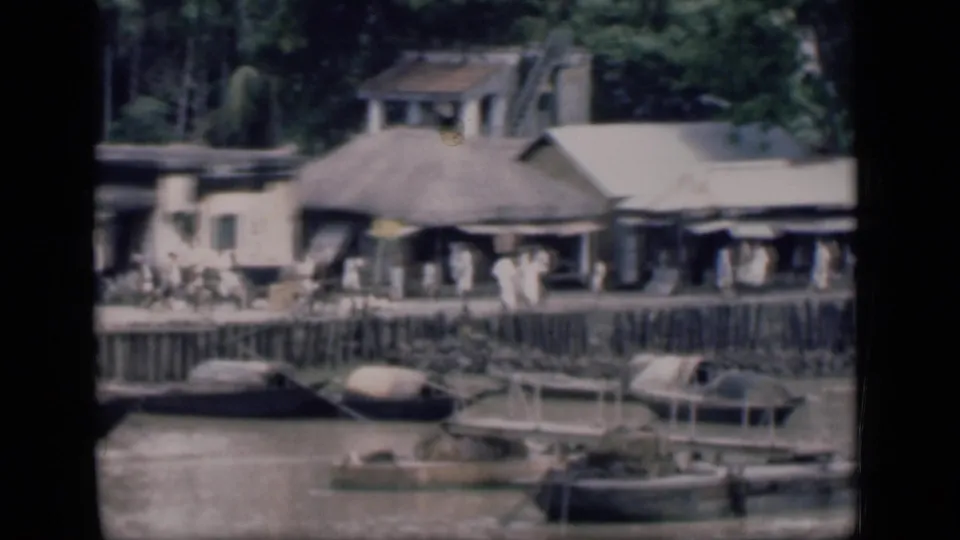 1946 Bombay home-movie frame shows a riverside settlement with low roofed structures, small boats, and people gathered along the waterline.