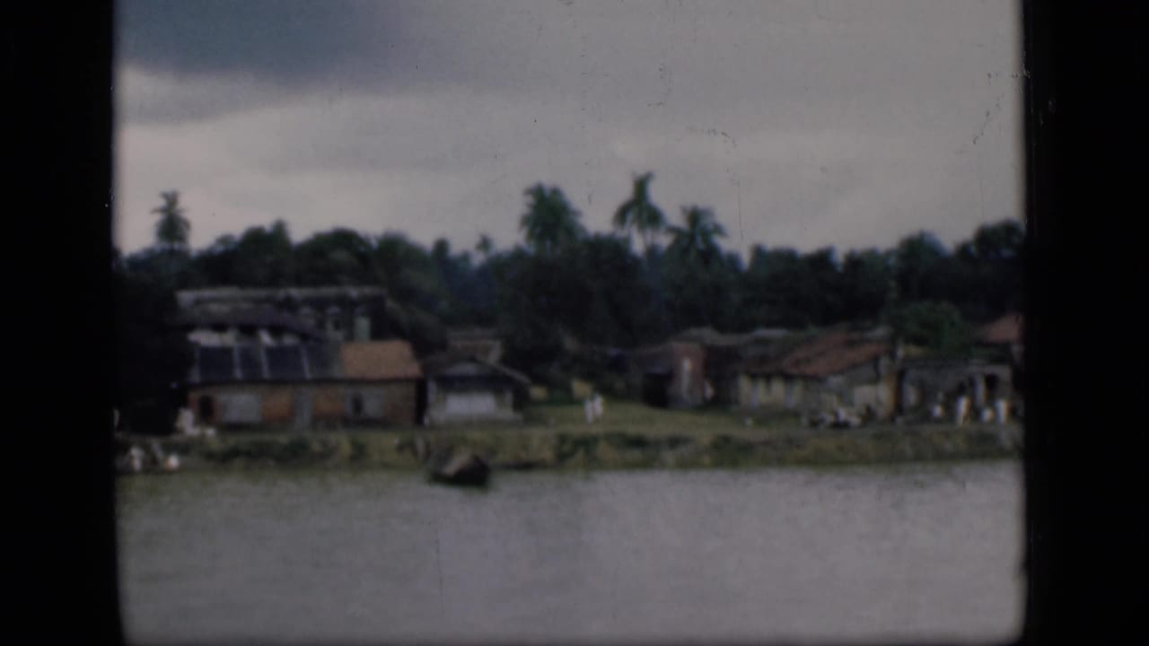 1946 Bombay riverside view with low homes, palm trees, and a small boat-like shape along the waterline.