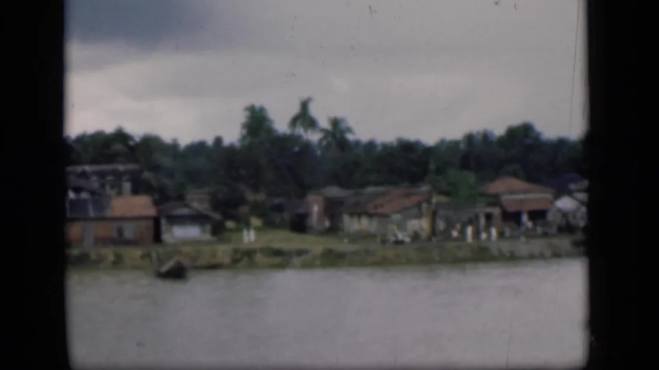 1946 Bombay home-movie frame of modest riverbank homes, palms, grass, and small distant people near the shoreline.