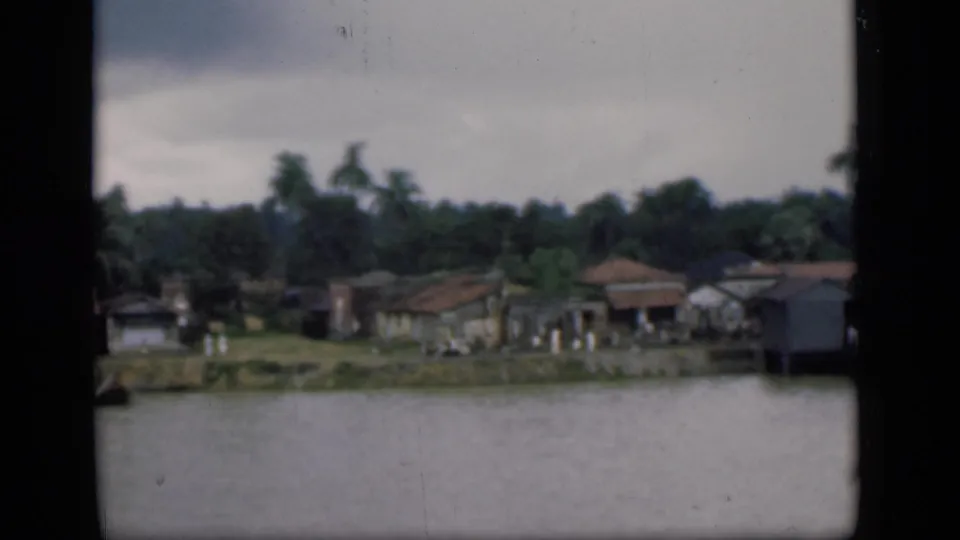 1946 Bombay riverside scene with clustered homes, palms, a grassy bank, and distant people beside the water.
